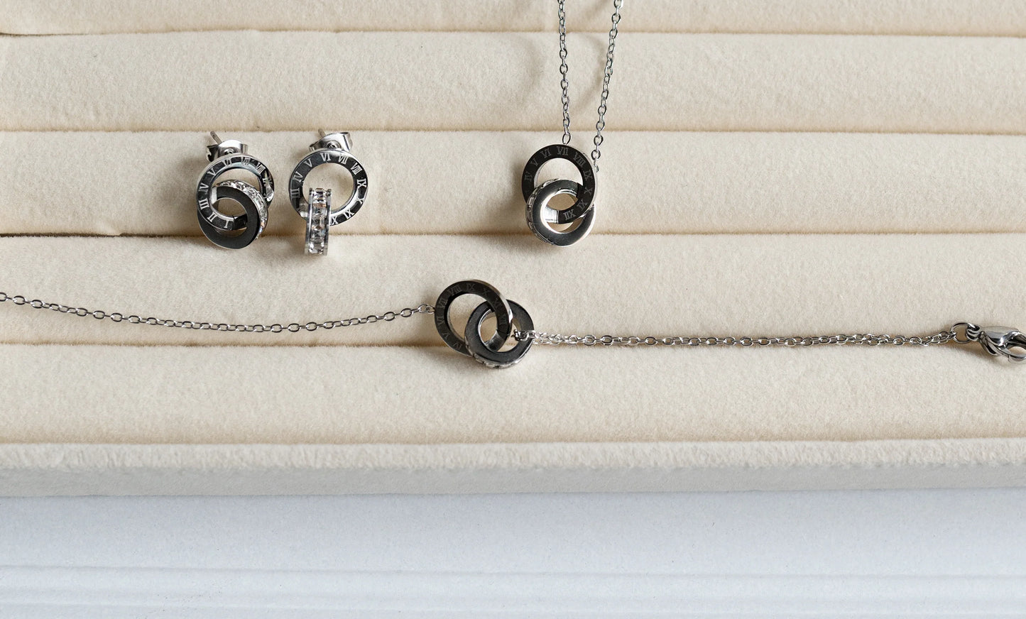 Set de bisutería fina MYKOANI con dijes entrelazados en acero, fondo beige elegante.