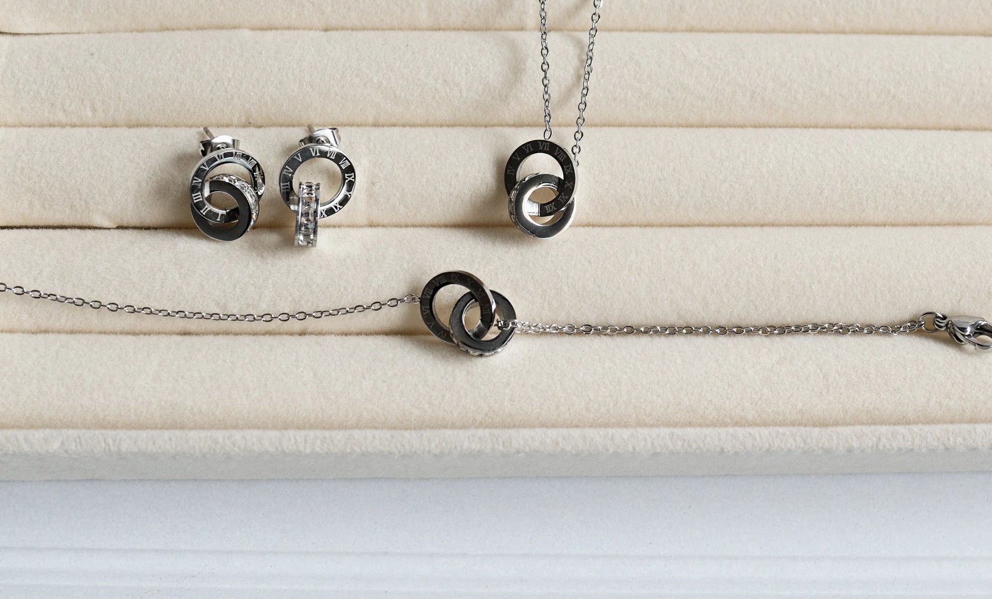 Set de bisutería fina MYKOANI con dijes entrelazados en acero, fondo beige elegante.