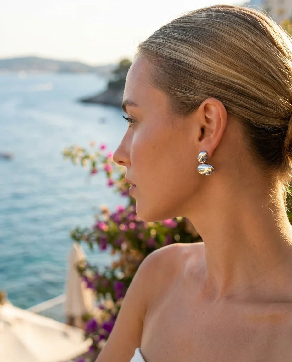 Mujer con aretes de alta bisutería MYKOANI, vista al mar y flores, estilo elegante y exclusivo.
